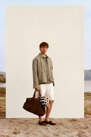 Male model wearing green jacket and white shorts holding a dark brown bag standing outdoors against a white panel on sand.