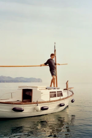 A man standing on the bow of a wooden boat on calm water under a pale sky