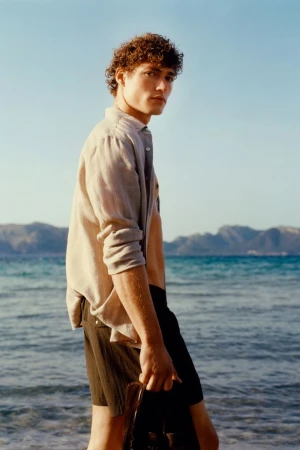 Model Valentin Humbroich wearing an open beige linen shirt and dark swim shorts standing by the sea on a sunny beach.