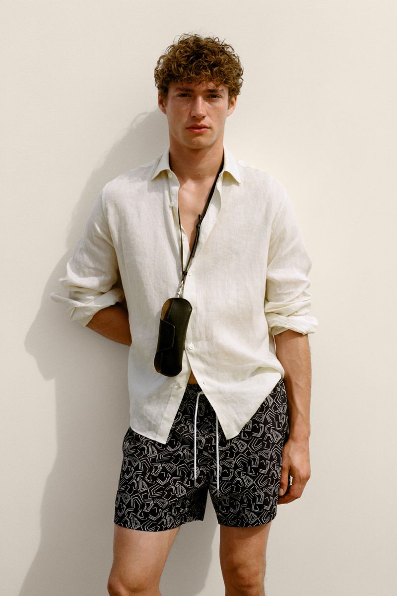 Model Valentin Humbroich wearing an open cream linen shirt and printed swim shorts against a plain white wall for Massimo Dutti.