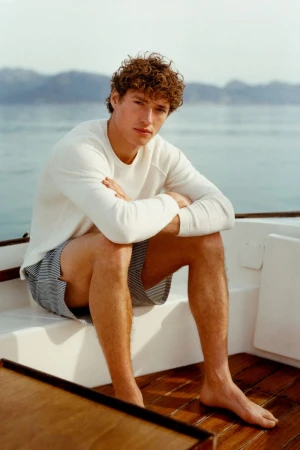 Model Valentin Humbroich wearing a white long-sleeve top and striped swim shorts seated barefoot on a boat deck.