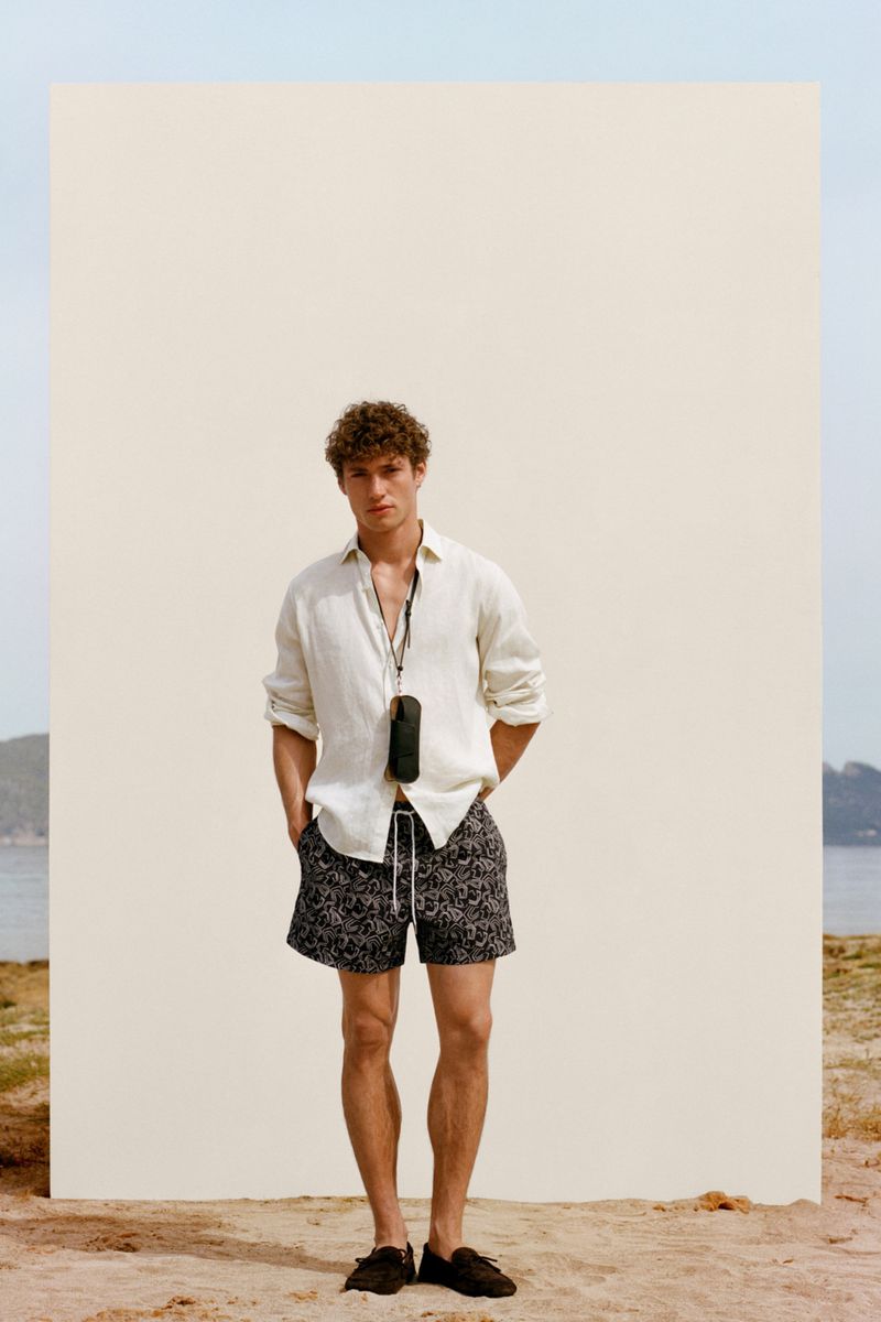 Model Valentin Humbroich in a white linen shirt and patterned swim shorts standing against a cream backdrop on a sandy beach.