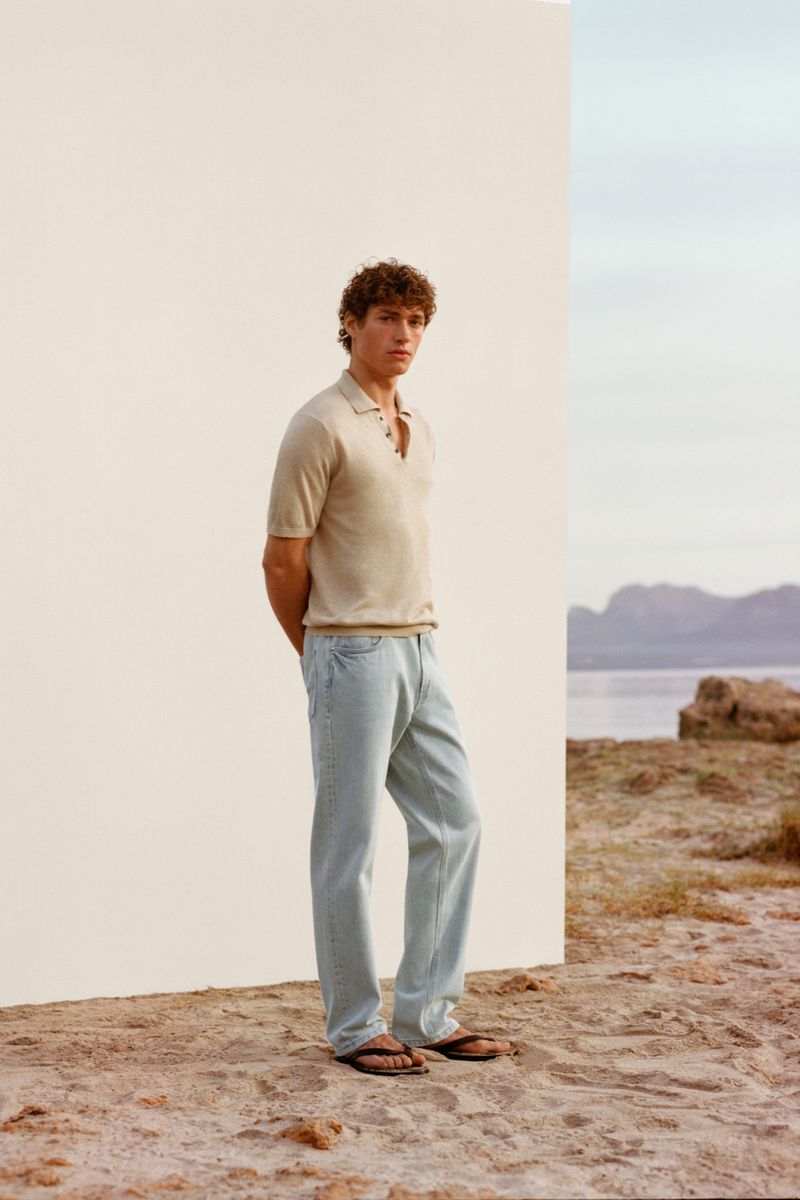 Model Valentin Humbroich poses in a beige knit polo shirt and light-wash jeans next to a white panel on a sandy beach.