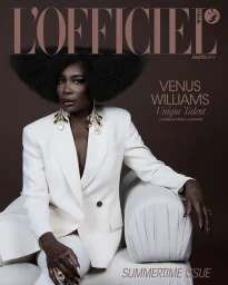 Venus Williams wearing a white tailored pantsuit and chandelier earrings seated on a modern white couch against a dark background.