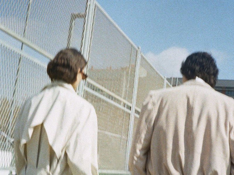 Rear view of two models wearing beige tailored coats walking beside a tall wire fence under daylight.