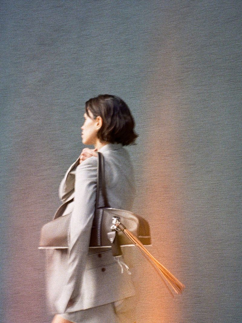 Model Noah Ramos carrying a brown leather shoulder bag with long fringe tassels, wearing a grey tailored blazer.