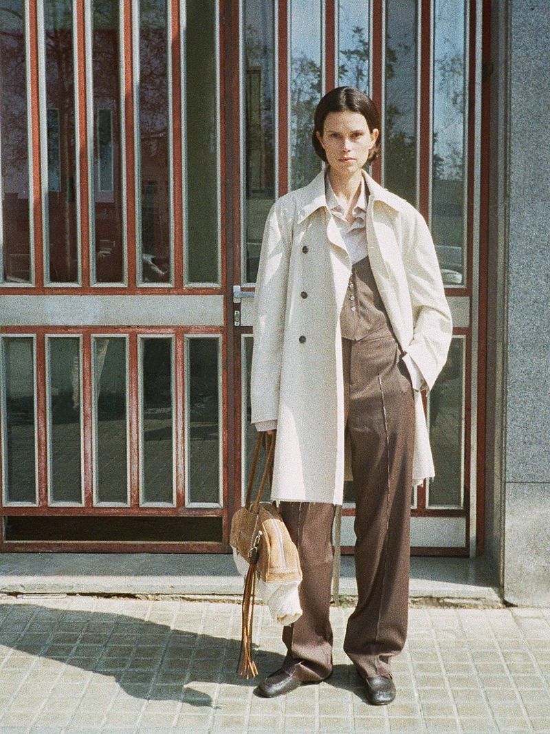 Model Noah Ramos wearing a cream trench coat and brown wide-leg trousers while holding a fringe handbag in front of a glass door.