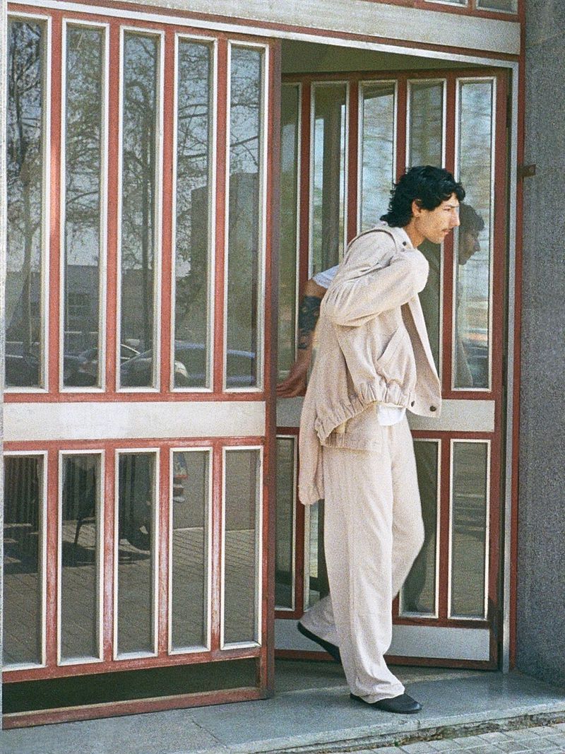Model Noah Ramos wearing a cream jacket and matching trousers while stepping through a red-framed glass doorway.
