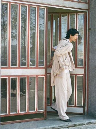 Model Noah Ramos wearing a cream jacket and matching trousers while stepping through a red-framed glass doorway.