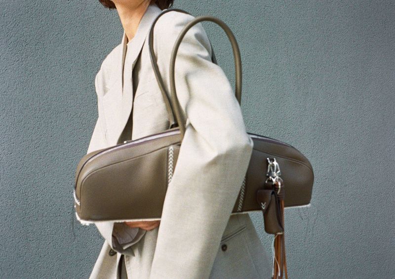 Model wearing a light grey blazer holding an elongated olive leather shoulder bag against a grey textured wall.