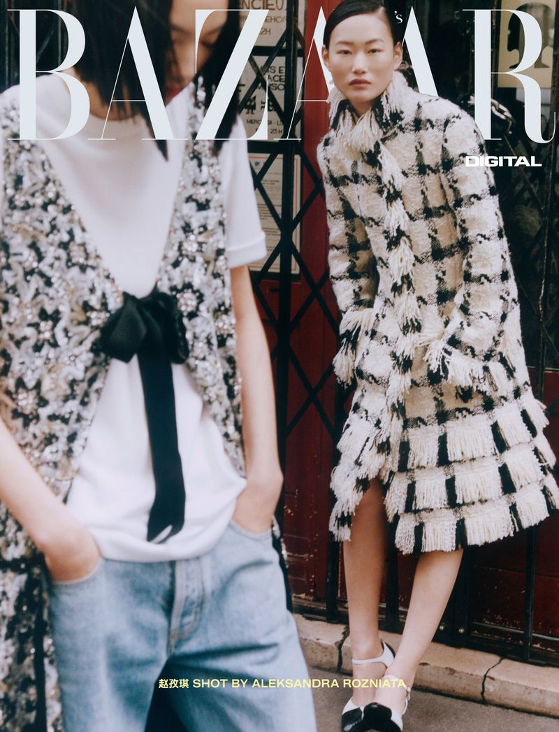 Two East Asian models posing in a street entrance, one wearing a fringed black-and-white plaid coat and the other in denim jeans.