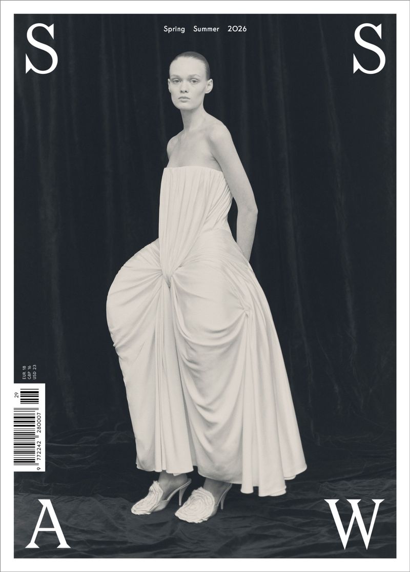 A model poses in a sculptural strapless white draped gown with exaggerated hips against a dark draped backdrop on a magazine cover.