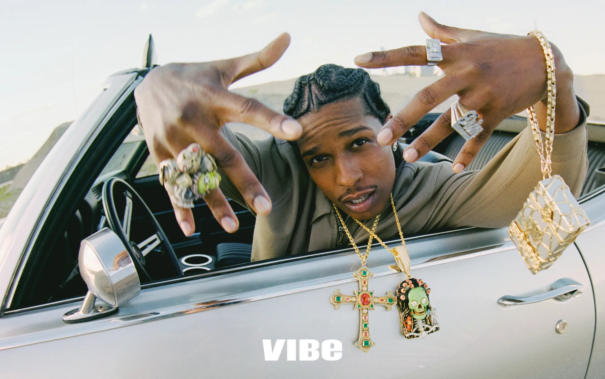 A$AP Rocky gesturing from inside a vintage convertible wearing layered gold jewelry and statement rings for Vibe Magazine.