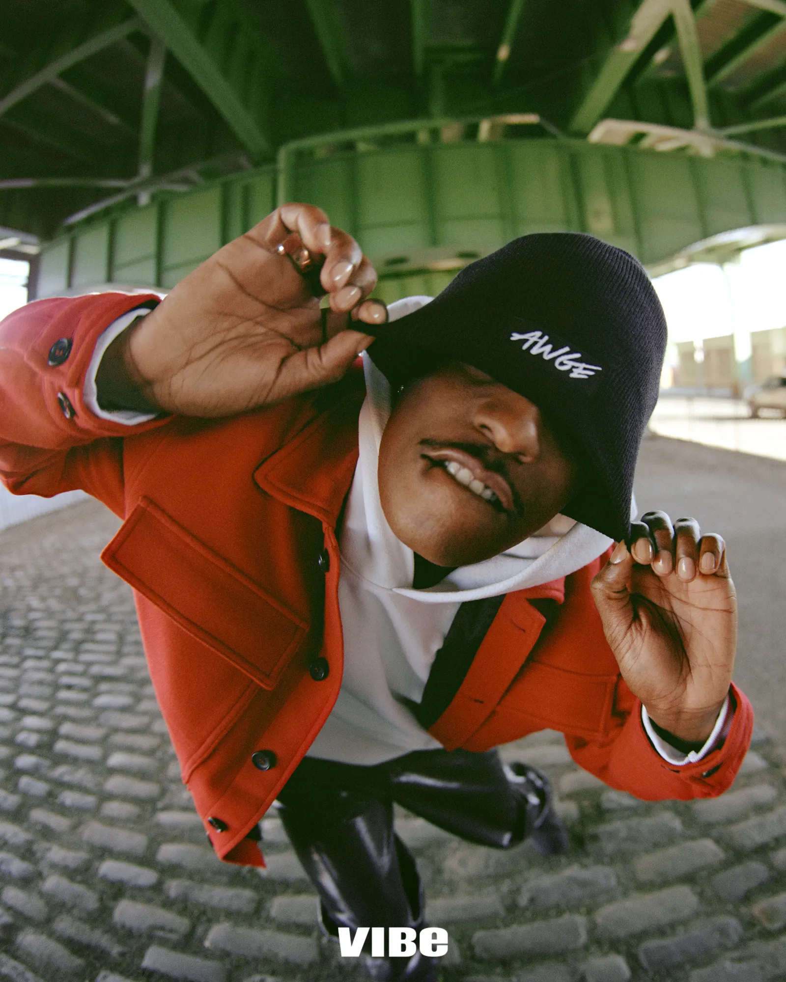 ASAP Rocky wearing a red jacket and black bucket hat outdoors beneath a green bridge