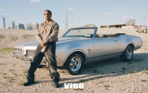 A$AP Rocky standing and leaning against a vintage silver convertible car on a gravel lot under clear daylight.