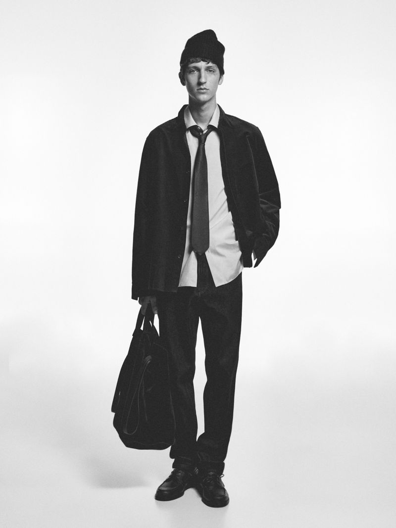 Model Saul Symon standing against a white backdrop wearing a black beanie, corduroy jacket, button-up shirt, tie, and holding a tote bag.