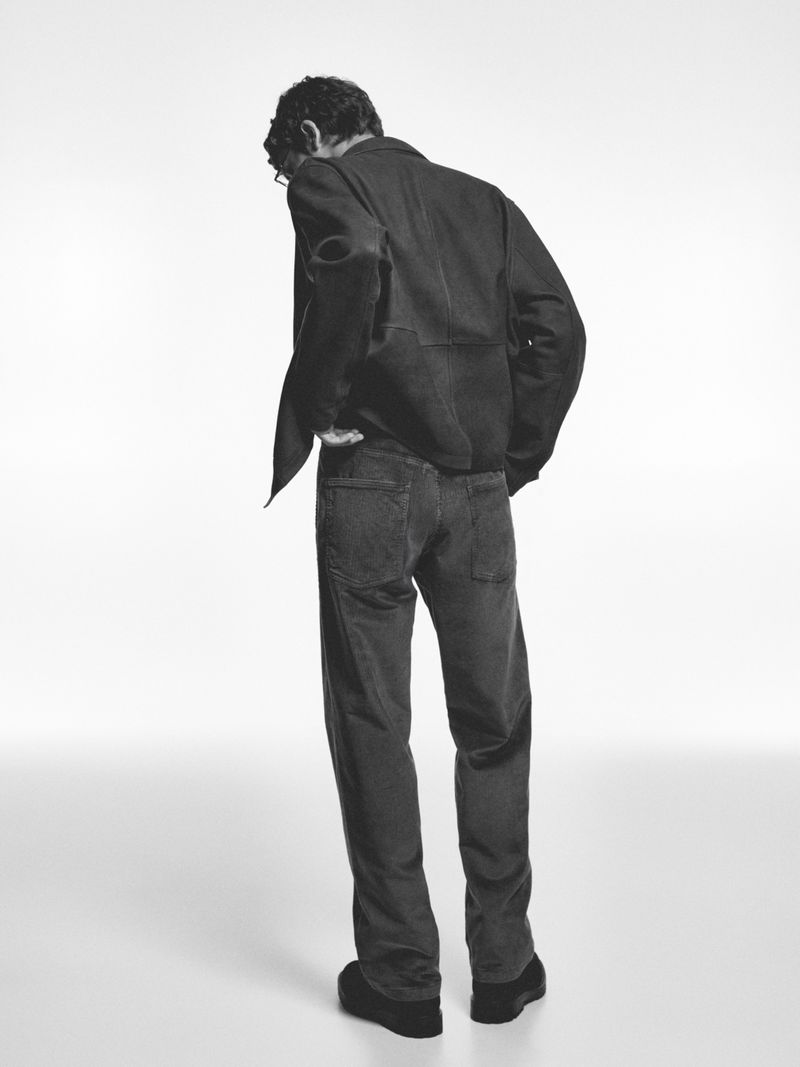 Full-length rear view of a male model wearing dark corduroy trousers and a jacket against a plain white studio background.