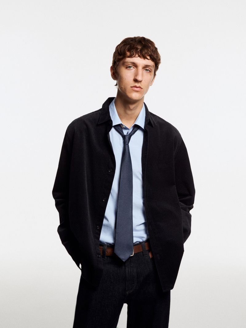 Model Saul Symon wearing a black corduroy overshirt, light blue shirt, navy tie, and dark jeans against a white seamless backdrop.