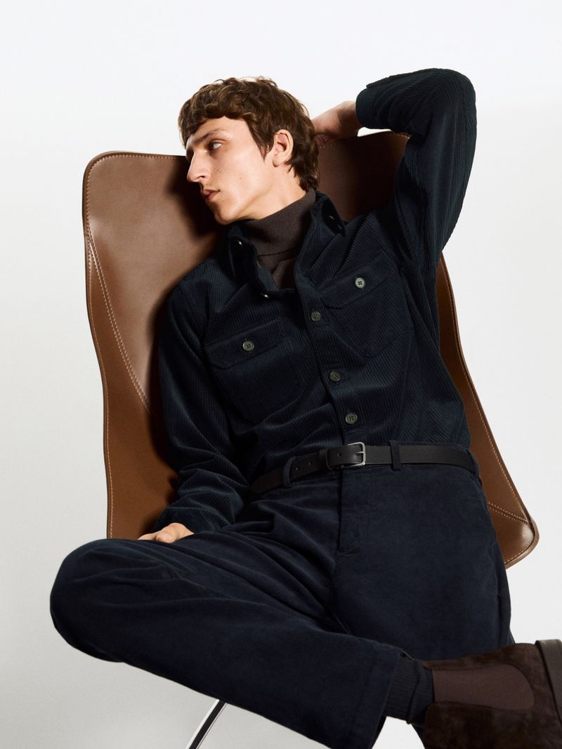 Model wearing dark corduroy shirt and trousers seated on a brown leather chair against a white background