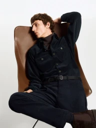 Model wearing dark corduroy shirt and trousers seated on a brown leather chair against a white background