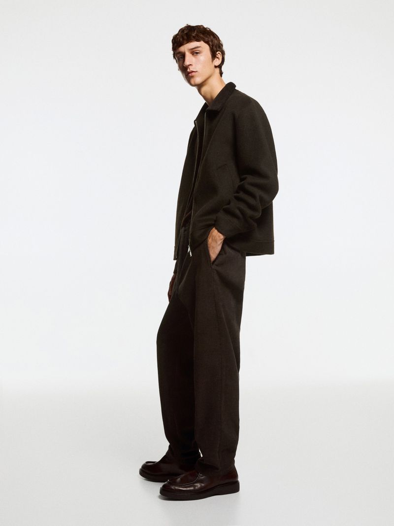 Model Saul Symon stands in a dark brown corduroy zip jacket and wide-leg trousers against a clean white seamless studio backdrop.