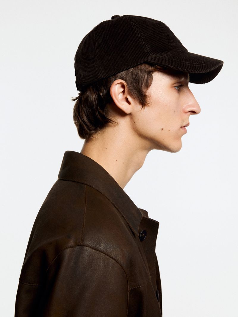 Profile shot of model Saul Symon wearing a dark corduroy baseball cap and brown jacket against a white studio background.