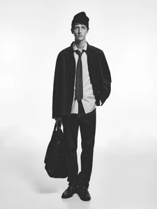 Model Saul Symon standing against a white backdrop wearing a black beanie, corduroy jacket, button-up shirt, tie, and holding a tote bag.