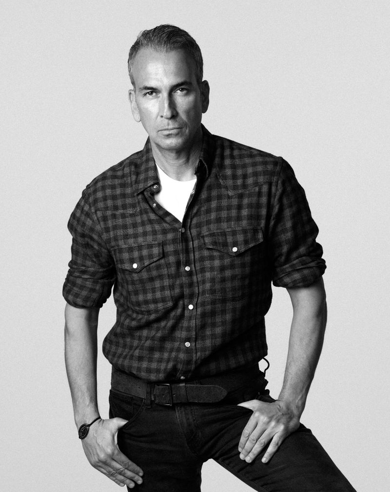 Black and white studio portrait of a man in a plaid button-down shirt and dark pants against a seamless grey backdrop.