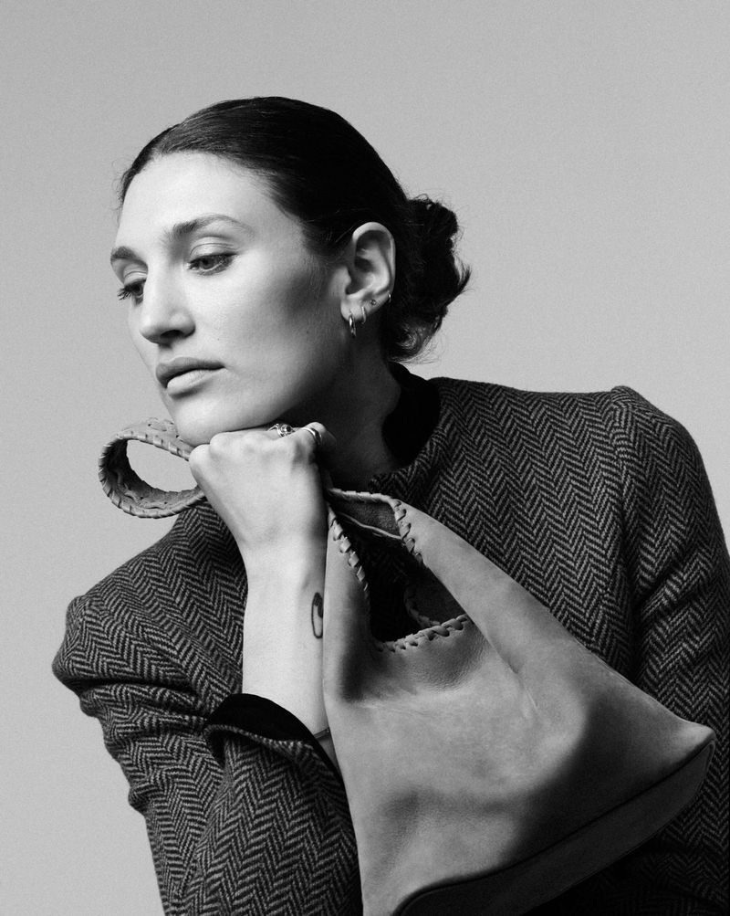 Black and white portrait of a woman in a herringbone jacket resting her chin on her hand while holding a structured leather bag.