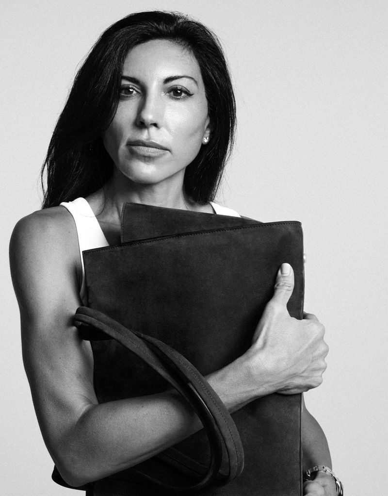 Black and white portrait of a dark-haired woman in a white tank top holding a large black tote bag against a plain background.