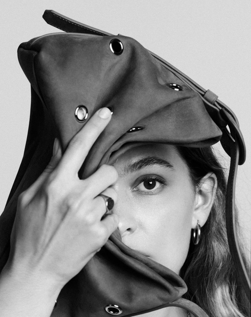 Close-up portrait of a woman holding a dark leather bag with grommets partly covering her face against a plain background