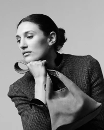 Black and white portrait of a woman in a herringbone jacket resting her chin on her hand while holding a structured leather bag.