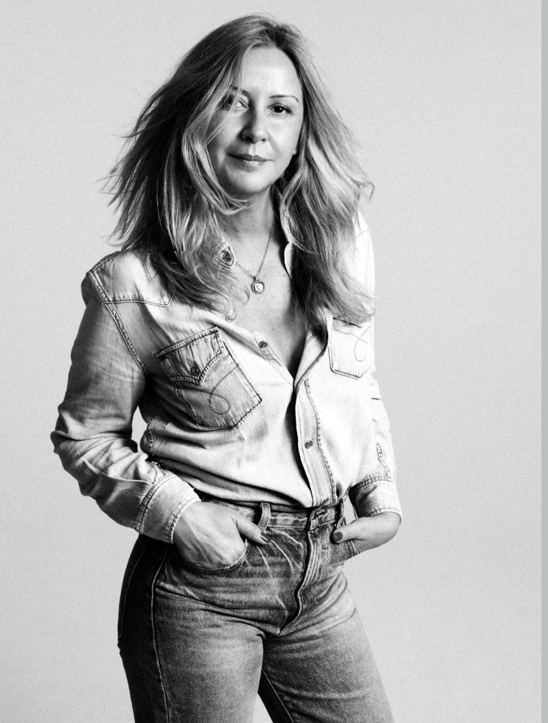 A woman with blonde layered hair wearing a denim shirt and jeans poses with hands in her pockets in black and white.