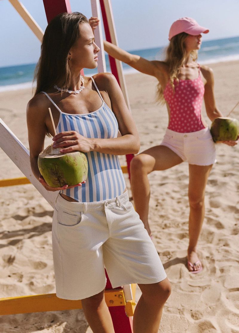 Two models holding coconuts wearing swimwear and shorts on a sunny sandy beach near a lifeguard tower.