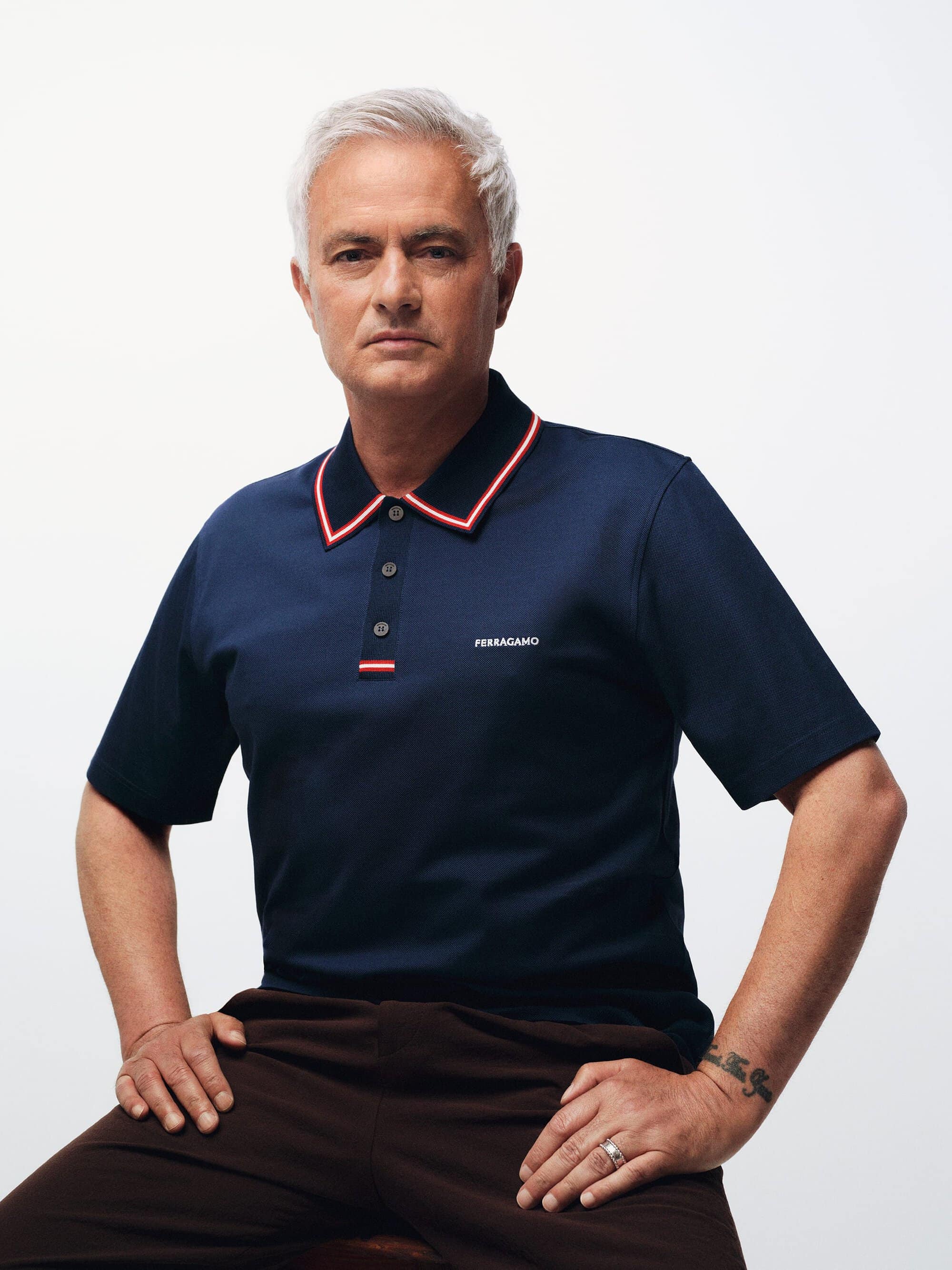 José Mourinho seated in a navy blue Ferragamo polo shirt with red collar trim against a white seamless background.