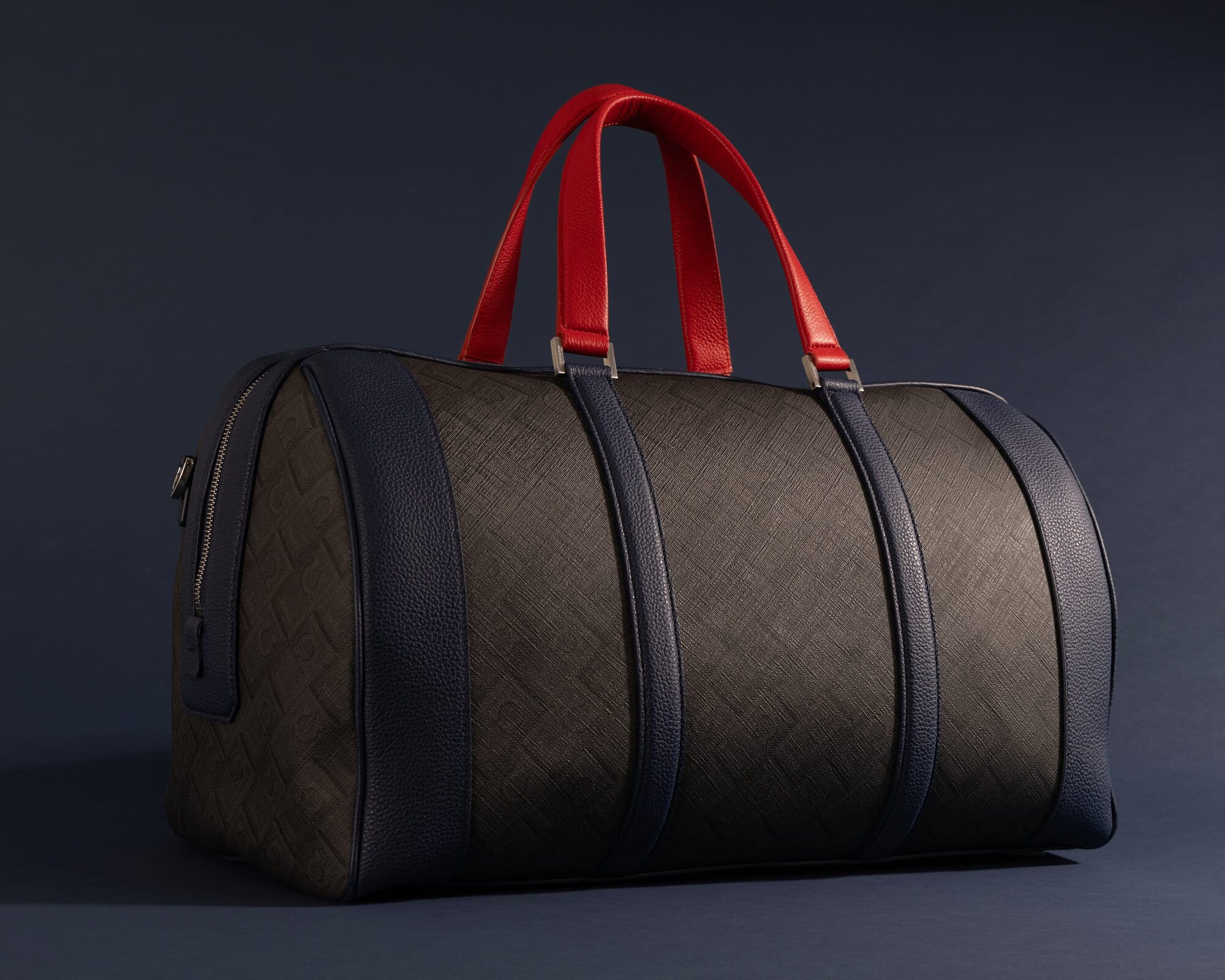 Ferragamo leather duffle bag with textured monogram body, dark navy trim, and vibrant red handles on a dark blue background.