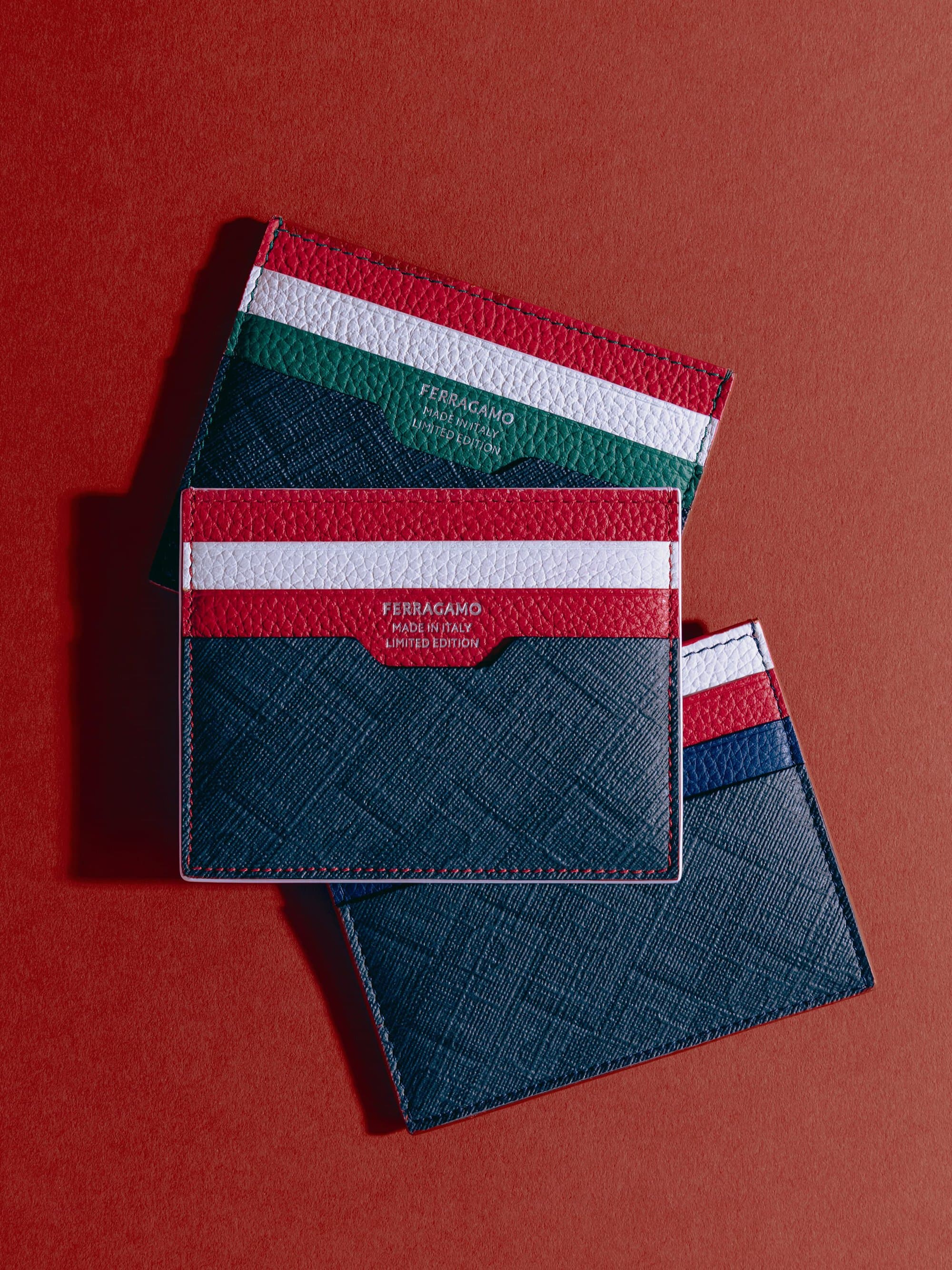 Three textured leather Ferragamo cardholders with colored stripes arranged flat against a solid red backdrop.