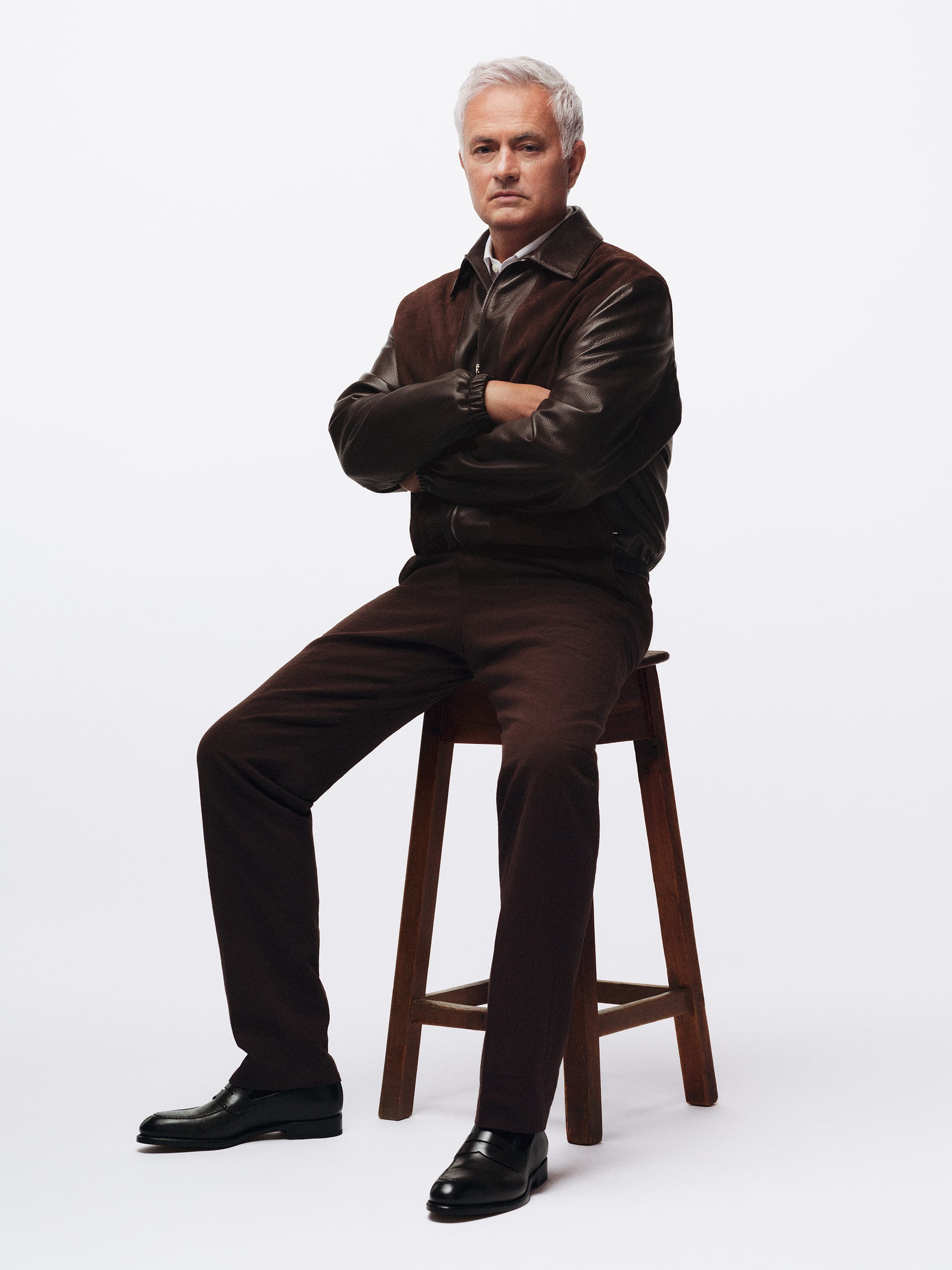 José Mourinho wearing a dark brown leather jacket and matching trousers seated on a wooden stool against a white seamless backdrop.