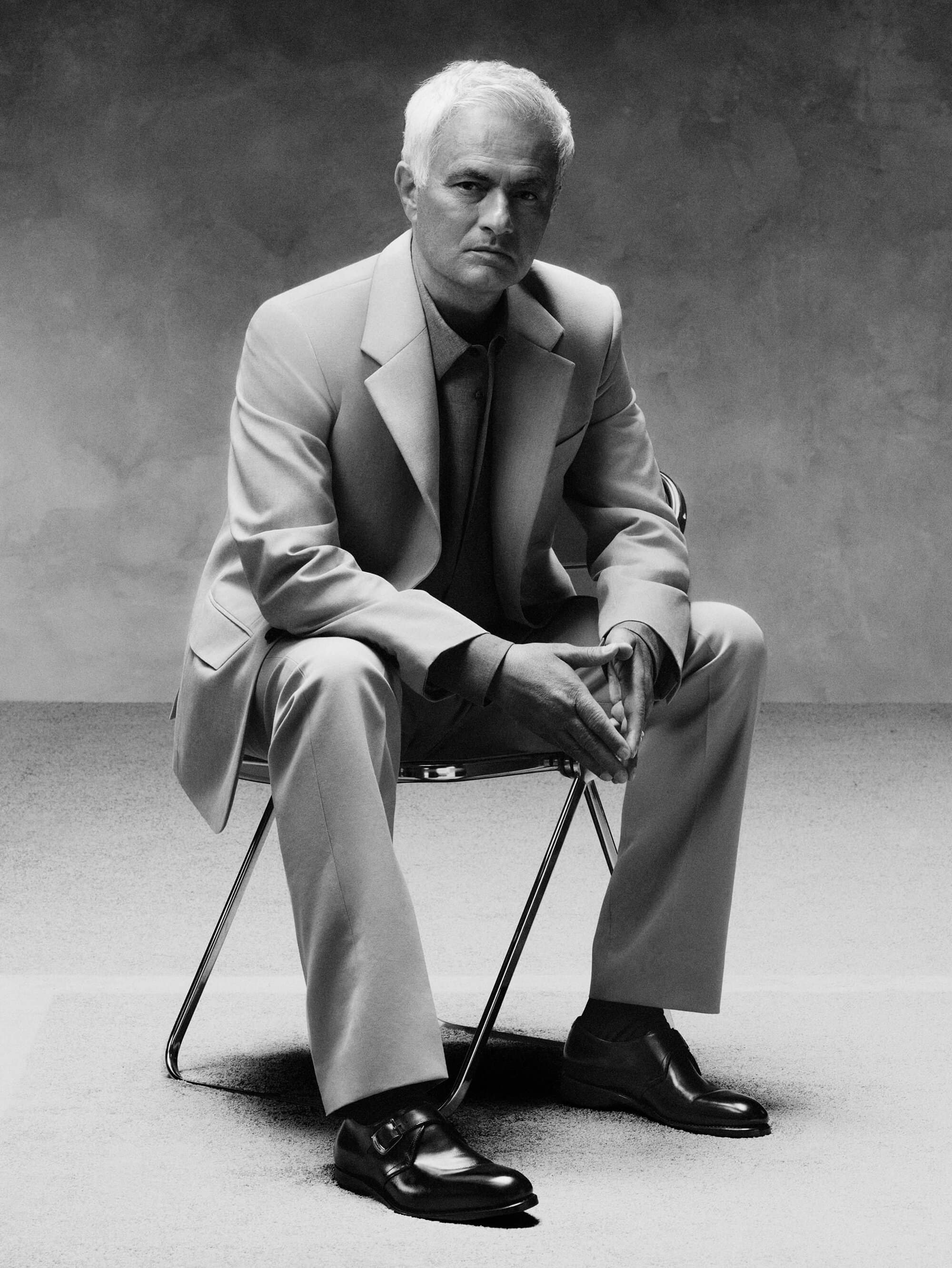 José Mourinho seated in a light tailored suit on a metal chair in a black and white studio portrait.