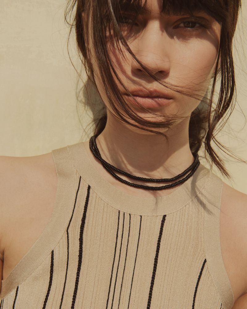 Close-up portrait of a woman wearing a beige knit top and a layered black beaded necklace against a plain wall.