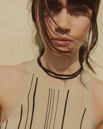 Close-up portrait of a woman wearing a beige knit top and a layered black beaded necklace against a plain wall.