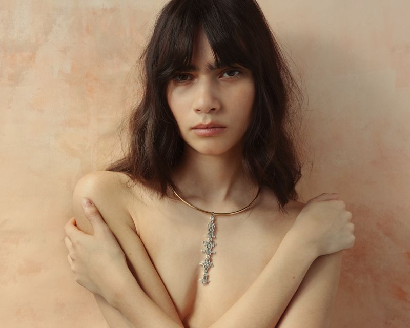 Model Clara Marques wearing a structured drop pendant choker necklace against a warm painterly textured background.