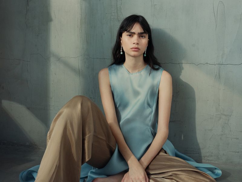Model seated on the floor wearing a pale blue sleeveless tunic and brown trousers against a weathered wall