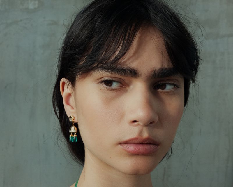 Close-up portrait of model Clara Marques wearing an ornate gold and green drop earring against a neutral grey textured wall.