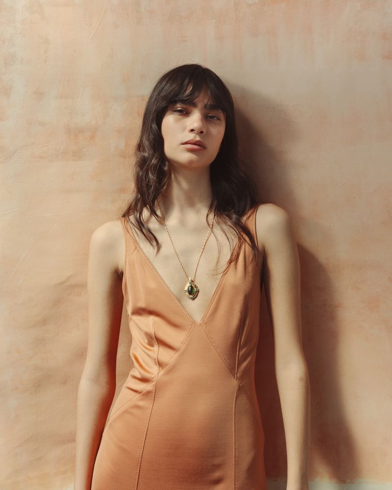 Model wearing a peach slip dress and pendant necklace posing against a warm textured wall.