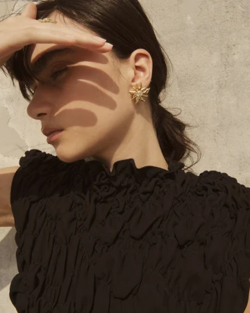 Model shielding her eyes in bright sunlight wearing a black ruched top and a statement gold sunburst earring against a textured wall.