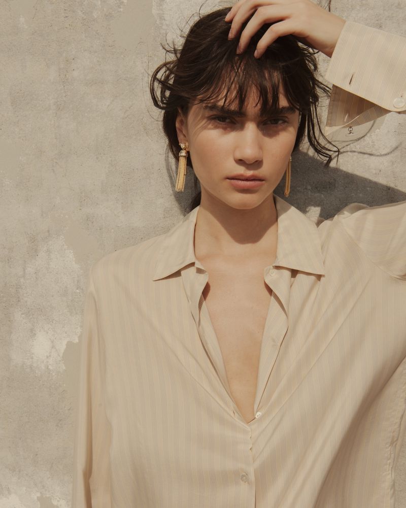 Clara Marques poses wearing gold tassel earrings and an unbuttoned cream shirt against a textured plaster wall.