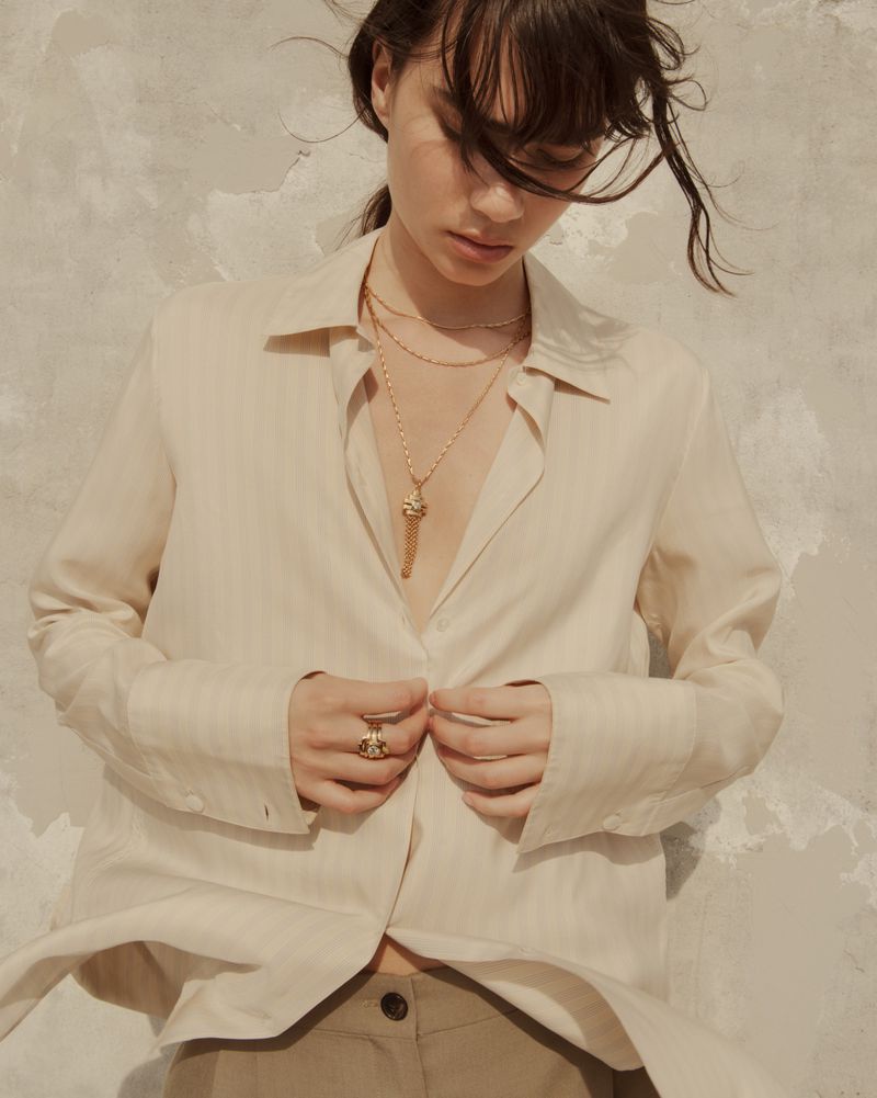 Model Clara Marques looking down while wearing layered gold necklaces and an oversized unbuttoned cream shirt.