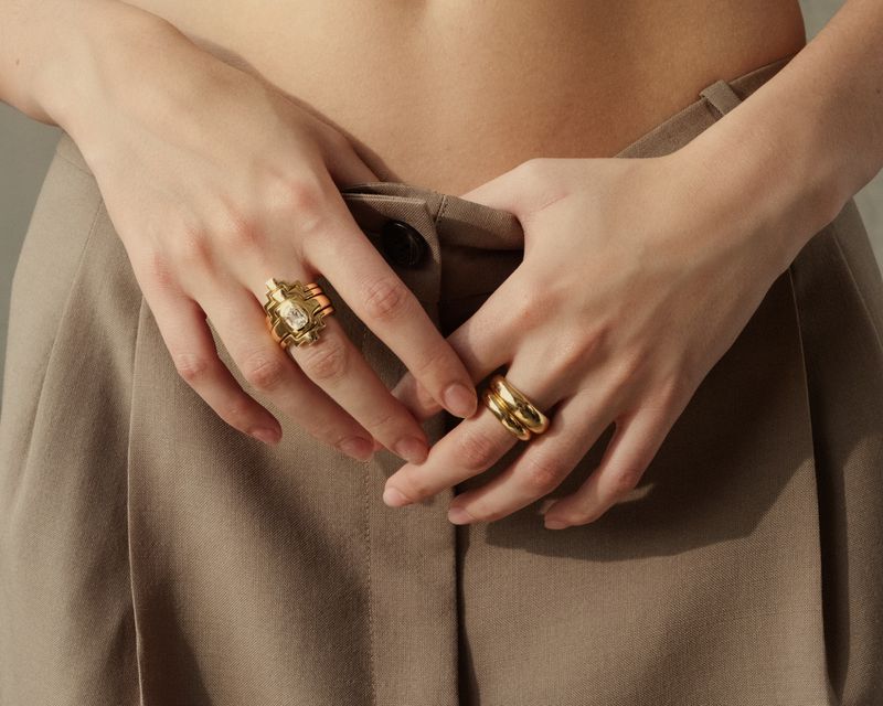 Close-up of model Clara Marques wearing gold rings on both hands against high-waisted beige pleated trousers.