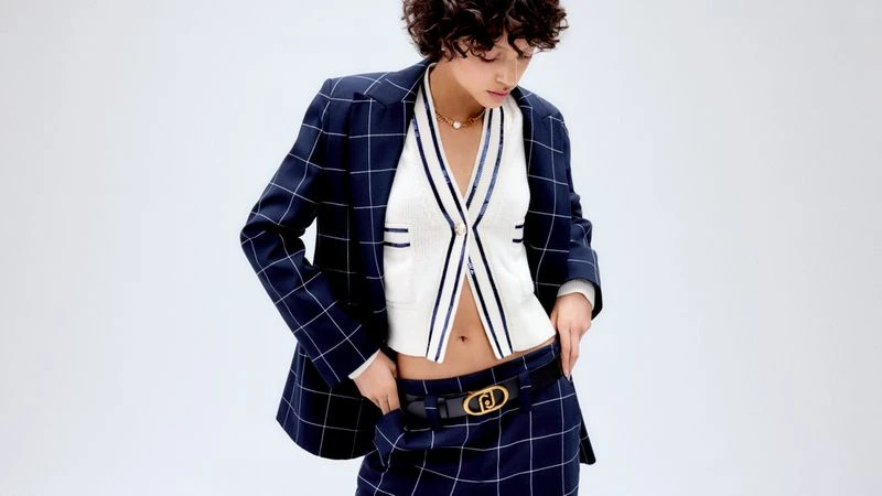 Woman in a navy plaid blazer and mini skirt with white cropped top and gold necklace poses in a studio with a light background.